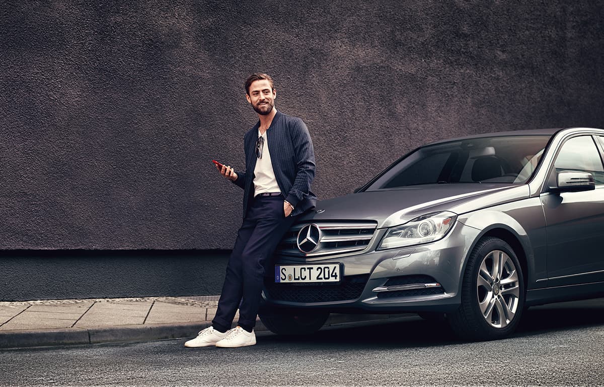 Ein Mann mit weißen Sneakern lehnt an der Motorhaube eines silbernen Mercedes-Benz Fahrzeugs.
