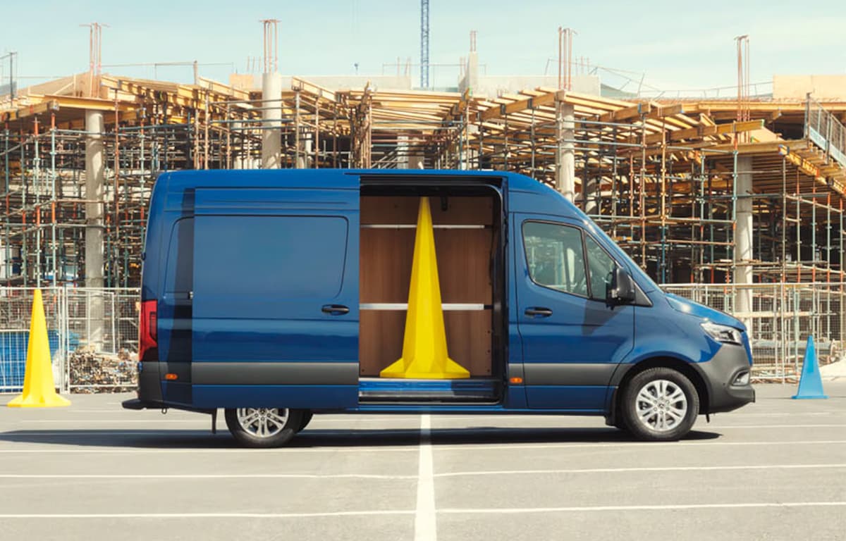Ein blauer Sprinter von der Seite mit geöffneter Schiebetür. Im Laderaum steht ein großer, stilisierter, gelber Pylone. Im Hintergrund eine Baustelle mit Gerüst.