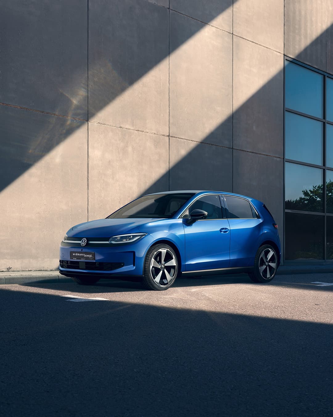 Ein blauer Kompaktwagen steht im Sonnenlicht vor einer modernen Betonwand, teilweise im Schatten.