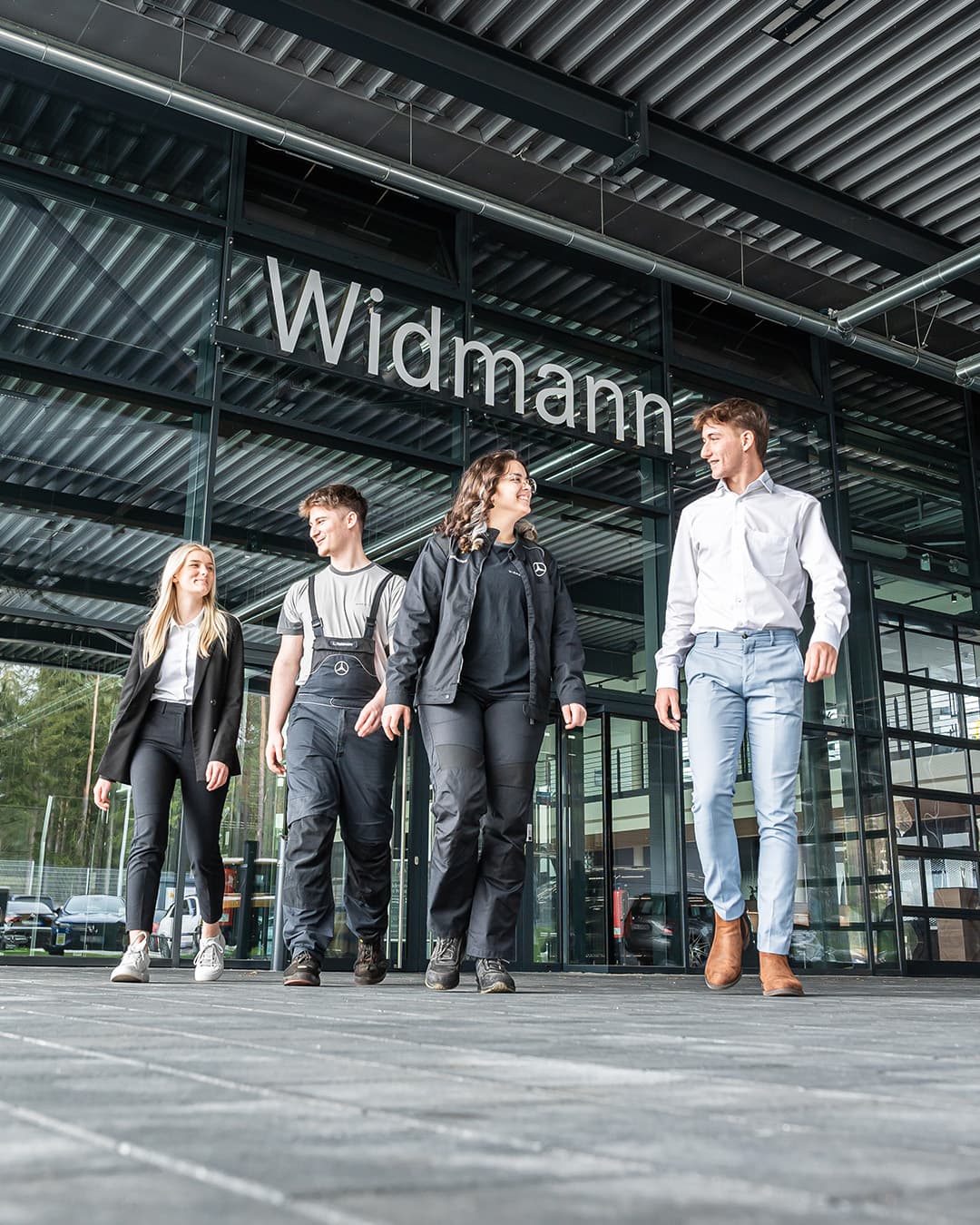 Vier junge Erwachsene aus unterschiedlichen Arbeitsbereichen gehen gemeinsam vor dem modernen, verglasten Eingangsbereich des Autohauses Widmann entlang.