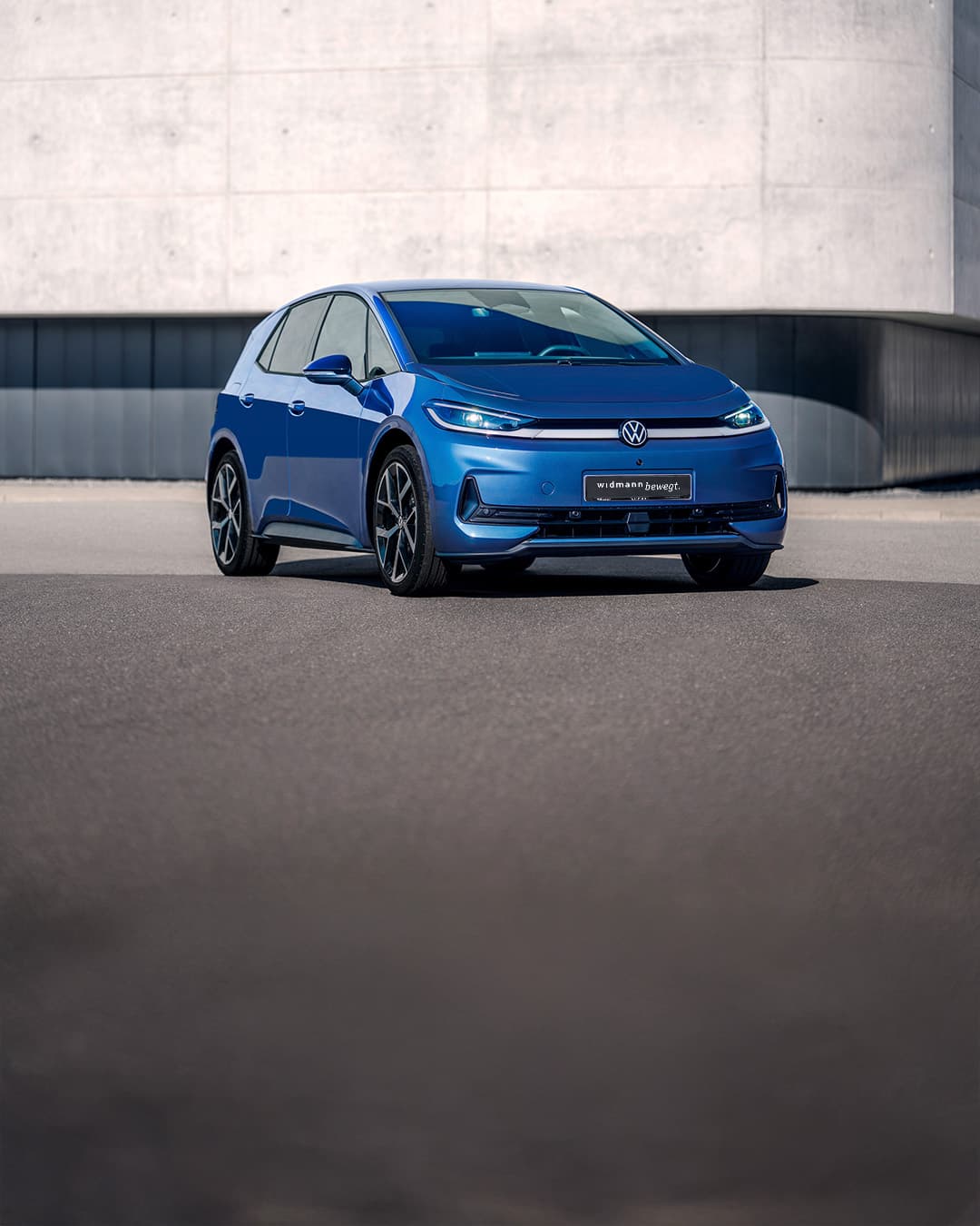 Ein blauer Volkswagen steht auf einer freien Fläche vor einer großen, schlichten Betonwand.