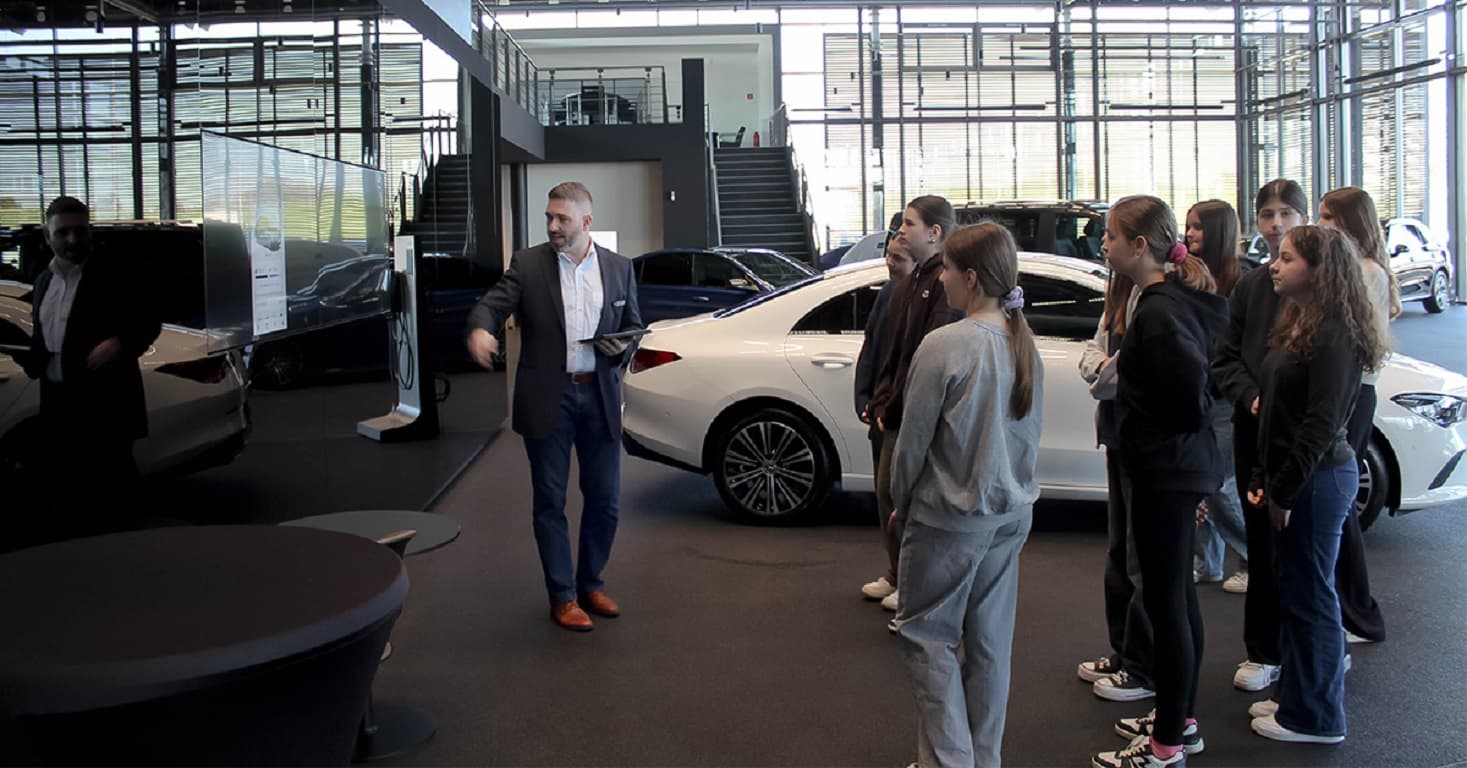 Ein Mann erklärt in einem Autohaus einer Gruppe junger Menschen ein Fahrzeug, während mehrere Autos im Showroom stehen.