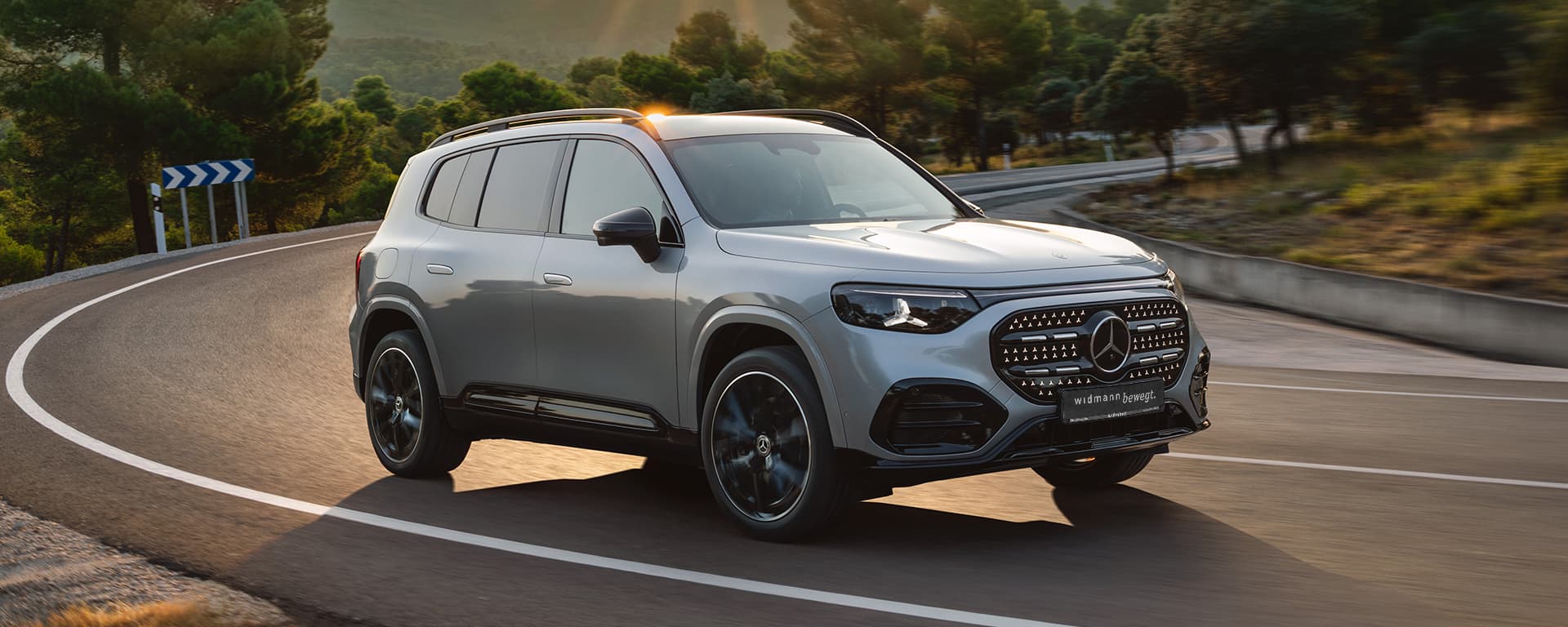 Ein silberner Mercedes-Benz GLB fährt auf einer kurvigen Straße durch eine grüne, bewaldete Landschaft im warmen Abendlicht.