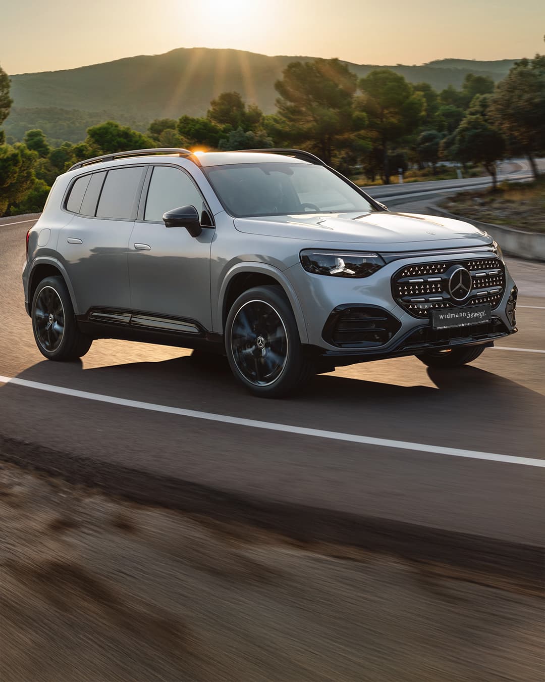 Ein silberner Mercedes-Benz GLB fährt auf einer kurvigen Straße durch eine grüne, bewaldete Landschaft im warmen Abendlicht.