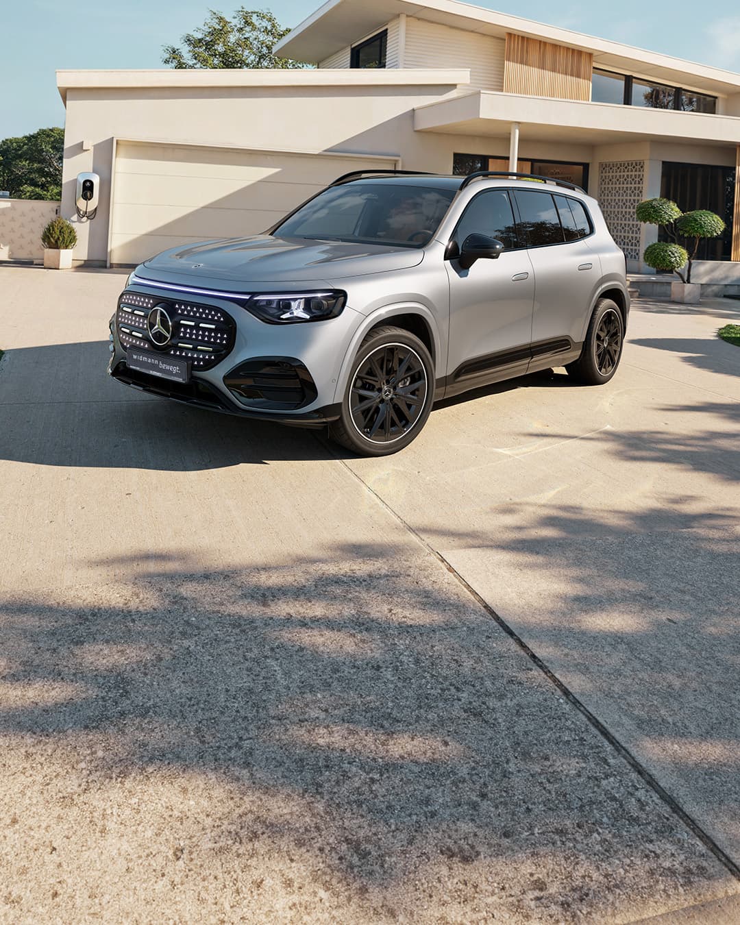 Ein silberner Mercedes-Benz GLB fährt auf einer kurvigen Straße durch eine grüne, bewaldete Landschaft im warmen Abendlicht.
