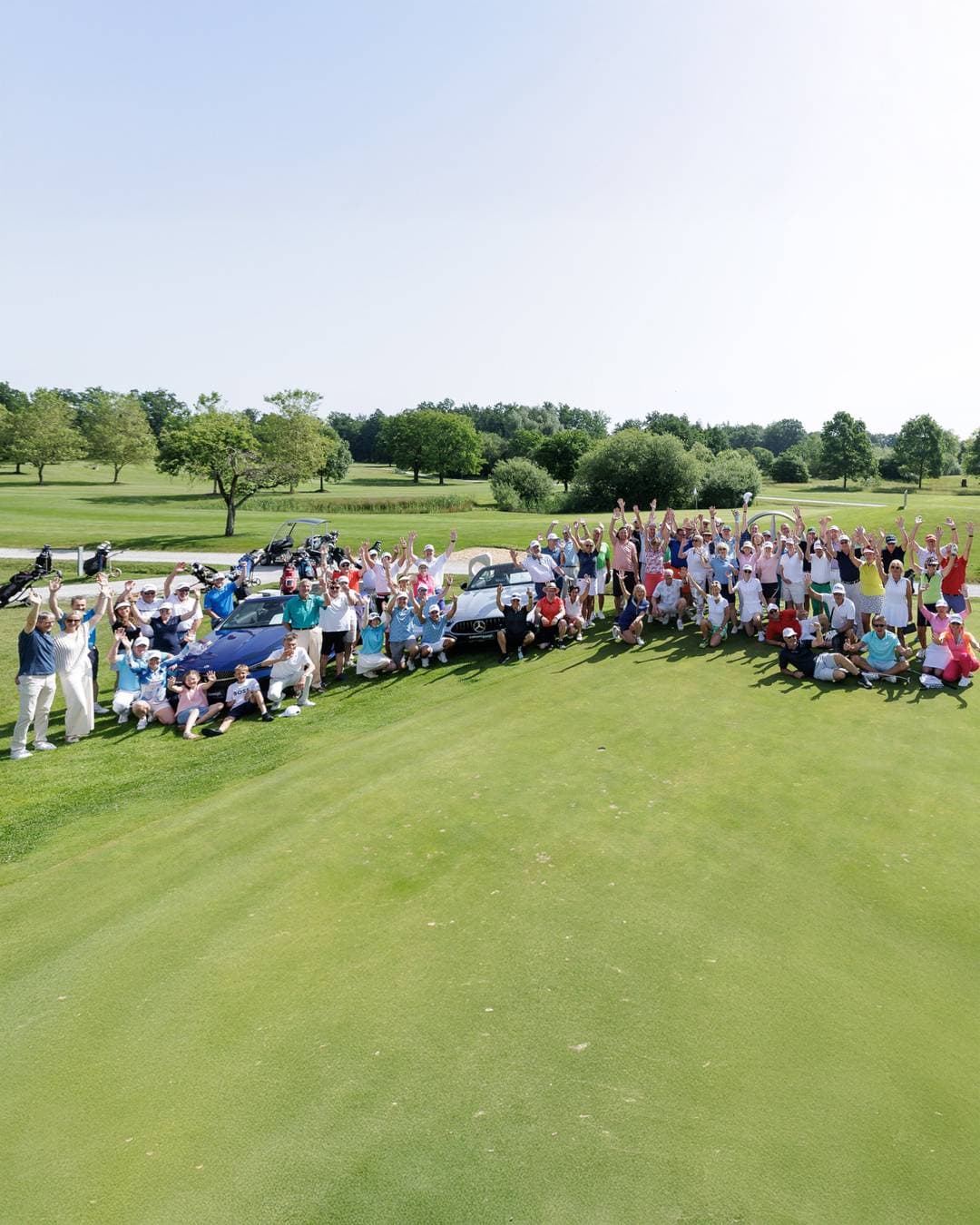 Die Teilnehmer eines Golfturniers in einem Halbkreis auf einem Golfplatz. Viele strecken die Arme in die Luft.