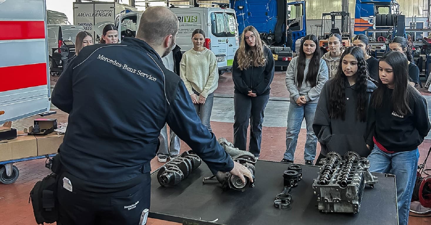 Ein Mechaniker erklärt in einer Werkhalle einer Gruppe junger Menschen an einem Tisch mit Motorbauteilen Fahrzeugtechnik.