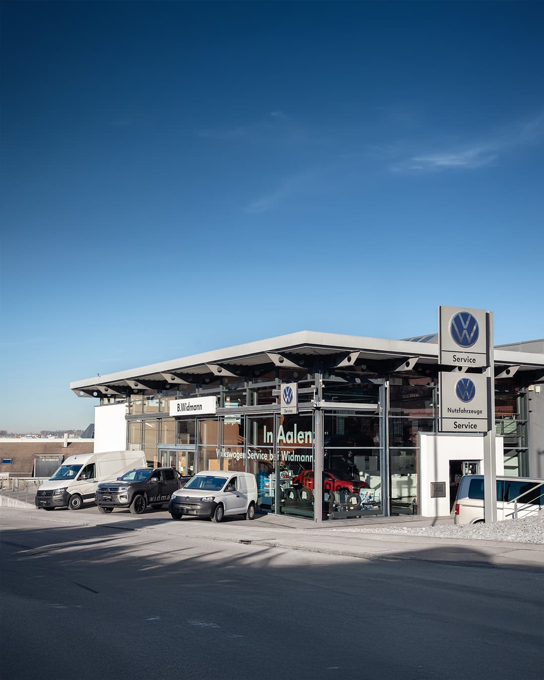 Ein modernes Autohaus mit VW Nutzfahrzeuge Service steht an einer Straße, vor dem mehrere Transporter geparkt sind.