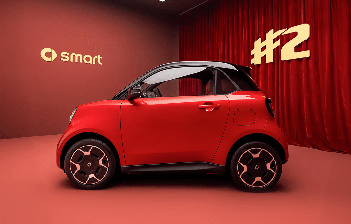 Roter smart #2 mit schwarzem Dach und markanten Leichtmetallfelgen in der Seitenansicht vor rotem Hintergrund mit goldenem smart-Logo und #2-Schriftzug.