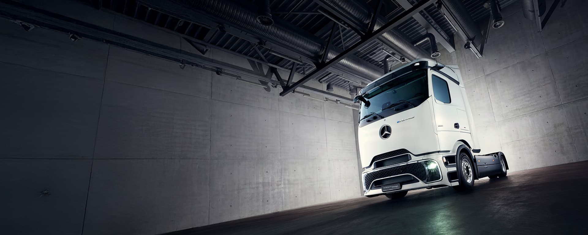 Ein weißer Mercedes-Benz Lkw steht in einer modernen, industriellen Halle mit Betonwänden und dunklem Boden, eindrucksvoll beleuchtet durch einen hellen Lichtkegel.