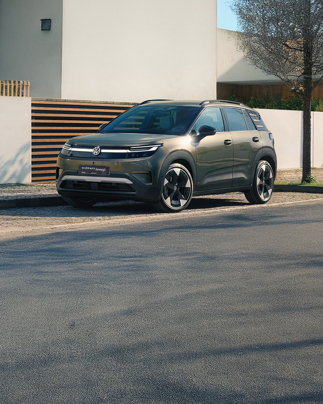Ein grüner Volkswagen ID. Cross steht vor einem modernen Wohnhaus mit heller Fassade, Holzelementen und Bäumen.