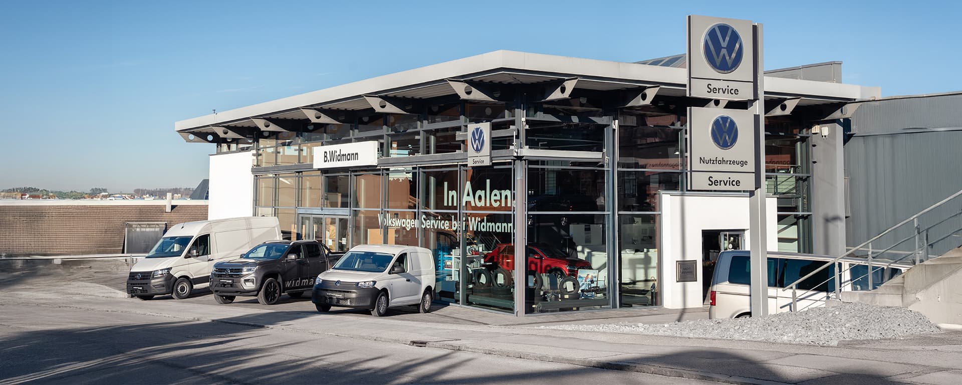 Ein modernes Autohaus mit VW Nutzfahrzeuge Service steht an einer Straße, vor dem mehrere Transporter geparkt sind.