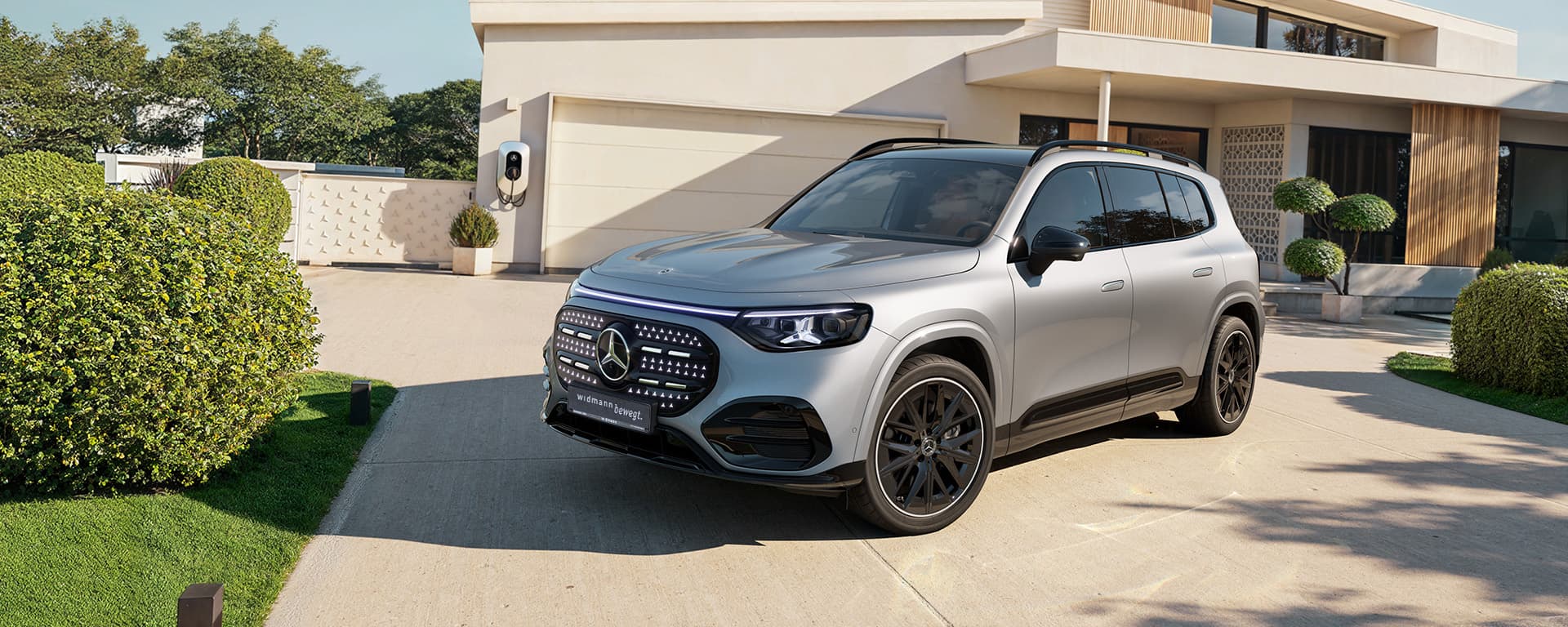 Ein silberner Mercedes-Benz GLB fährt auf einer kurvigen Straße durch eine grüne, bewaldete Landschaft im warmen Abendlicht.