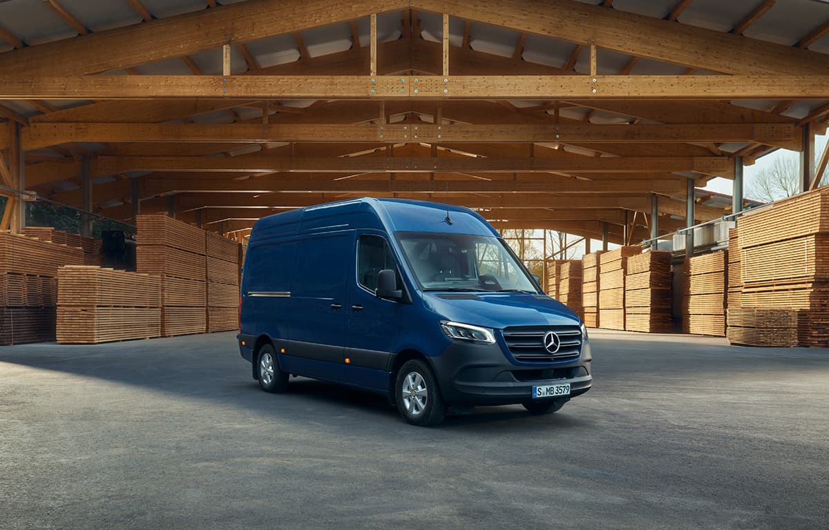 Ein dunkelblauer Mercedes-Benz Transporter steht in einer großen, offenen Holzlagerhalle mit gestapelten Holzbrettern entlang der Seitenwände.