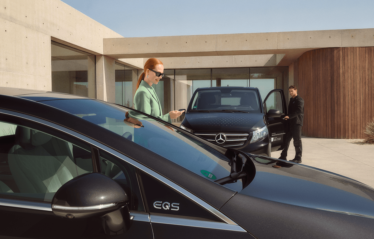 Eine Frau steht neben einem schwarzen Mercedes und schaut auf ihr Smartphone, während ein Techniker im Hintergrund an einem Servicefahrzeug arbeitet