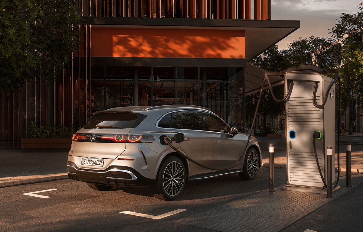 Ein silberner Mercedes kompakter SUV lädt an einer öffentlichen Ladesäule vor einem modernen Gebäude im warmen Abendlicht.