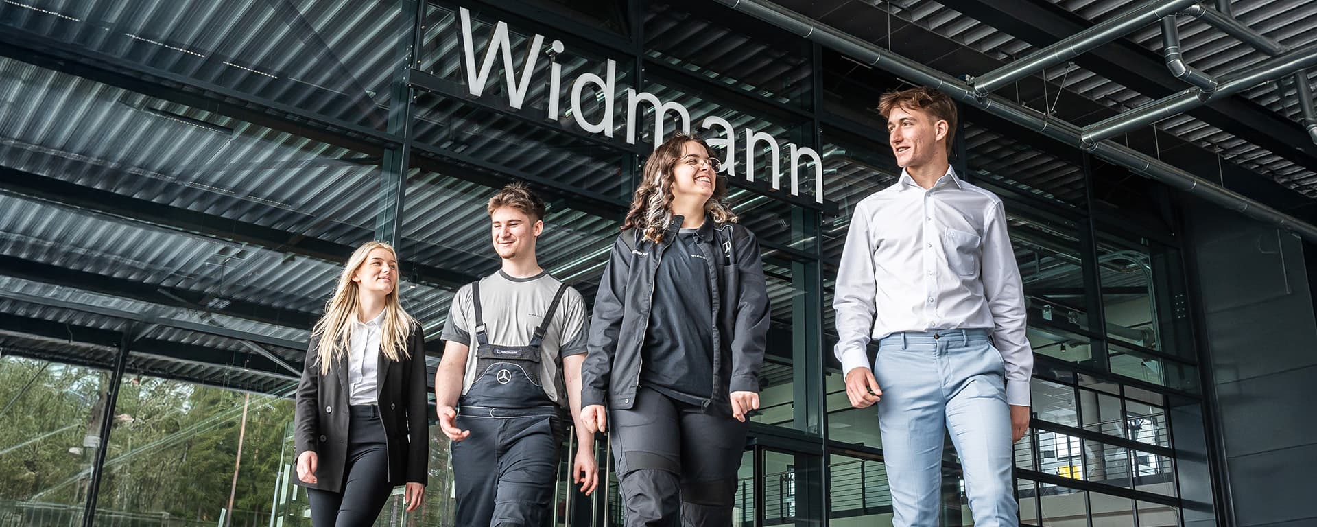 Vier junge Erwachsene aus unterschiedlichen Arbeitsbereichen gehen gemeinsam vor dem modernen, verglasten Eingangsbereich des Autohauses Widmann entlang.