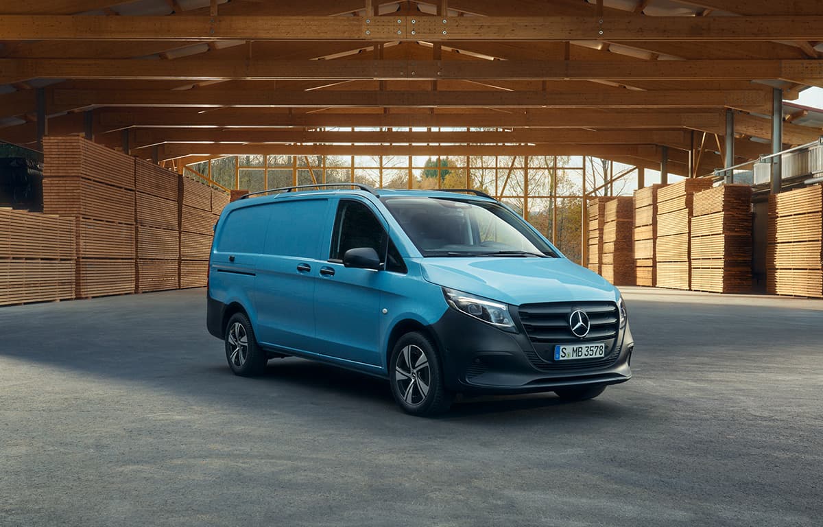 Ein hellblauer Mercedes-Benz Transporter steht in einer großen Holzlagerhalle mit hohen Dachbalken und gestapelten Holzbrettern im Hintergrund.