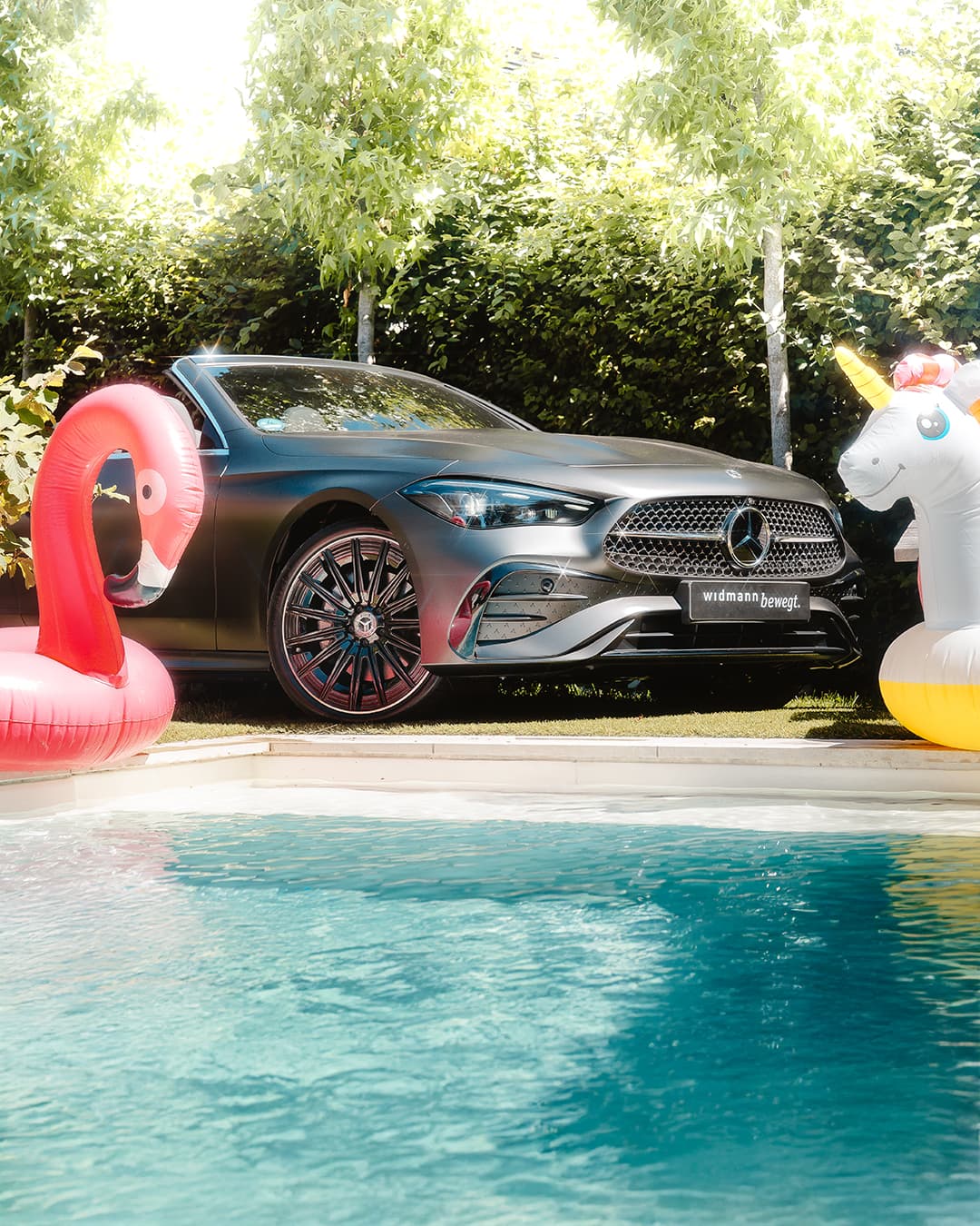 Ein aufblasbarer pinker Flamingo-Schwimmreif und ein aufblasbarer weißer Einhorn-Schwimmreif in einem Swimming Pool im Vordergrund. Im Hintergrund ein mattgraues Mercedes-Benz Cabrio.