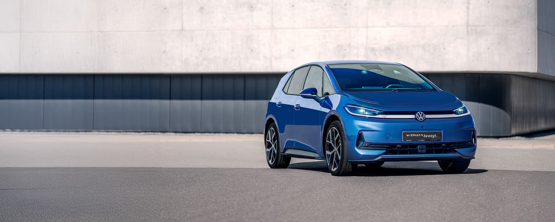 Ein blauer Volkswagen steht auf einer freien Fläche vor einer großen, schlichten Betonwand.