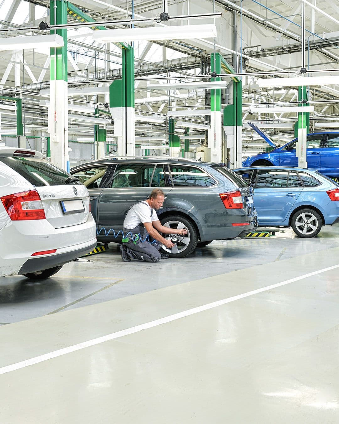 Mehrere Fahrzeuge stehen in einer großen Werkstatt, während ein Mechaniker an einem Auto arbeitet und andere Fahrzeuge auf Hebebühnen gewartet werden