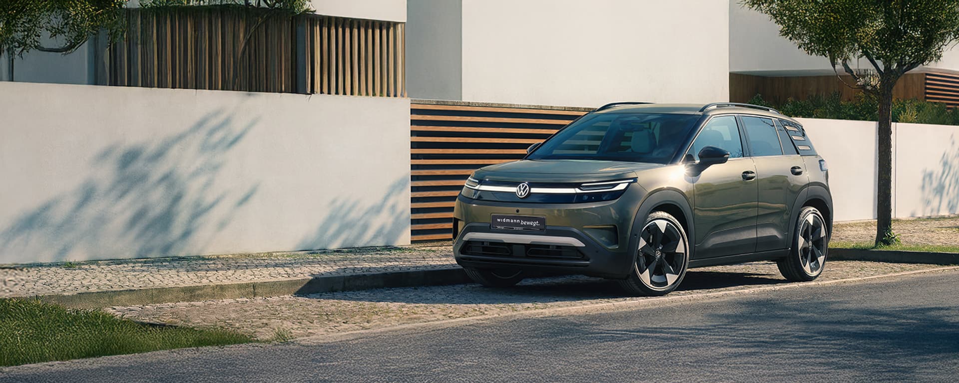Ein grüner Volkswagen ID. Cross steht vor einem modernen Wohnhaus mit heller Fassade, Holzelementen und Bäumen.