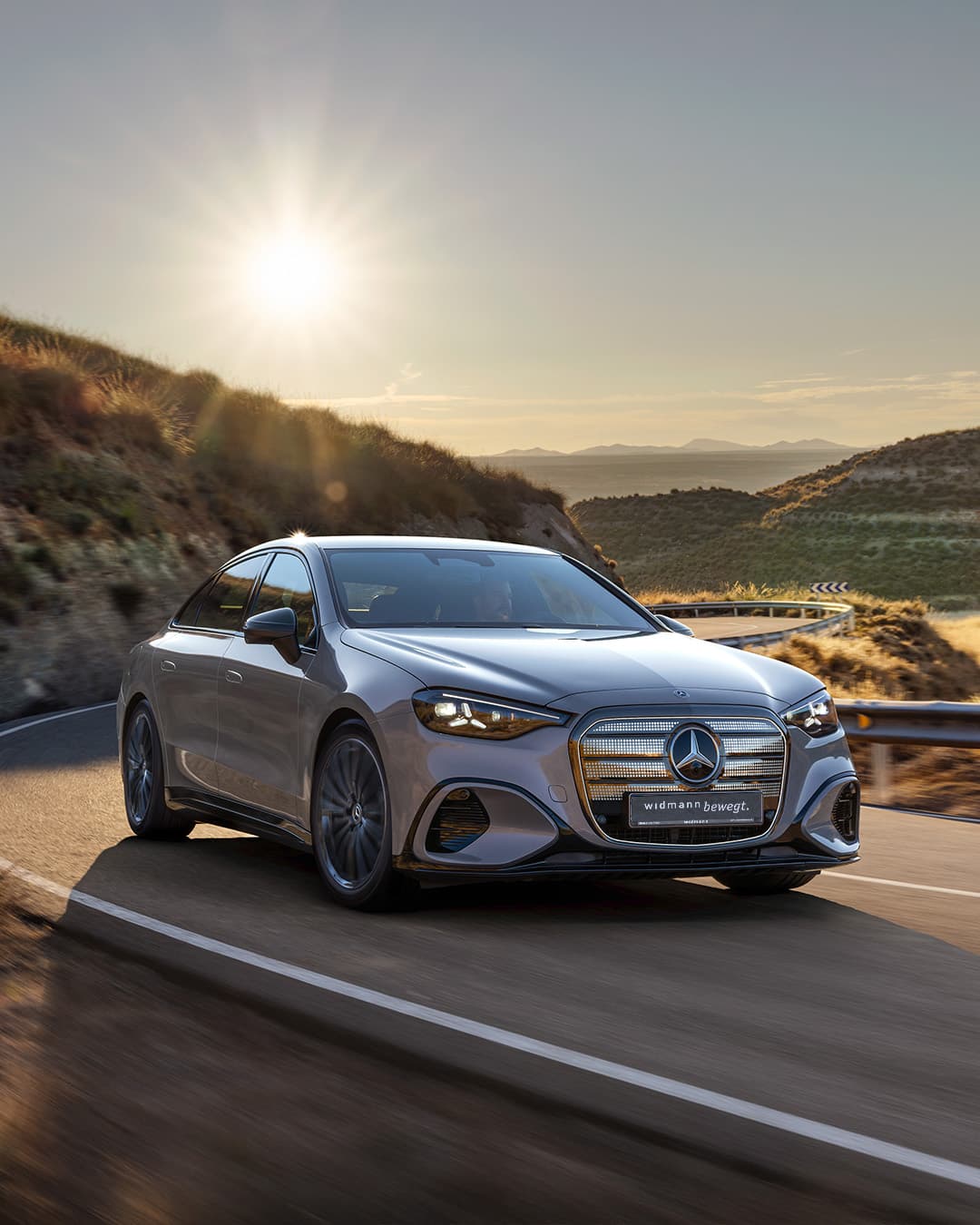 Ein silbernes Auto fährt auf einer kurvigen Straße durch eine hügelige Landschaft bei tief stehender Sonne.
