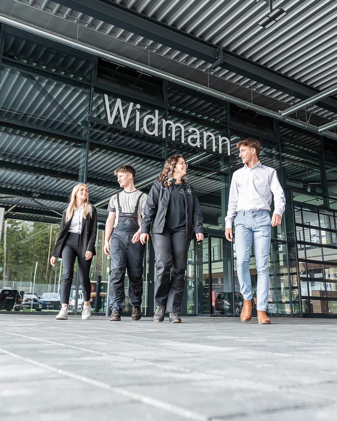 Fünf Mitarbeitende gehen gemeinsam vor dem modernen Glasgebäude von Widmann, dessen Name gut sichtbar über dem Eingang steht.