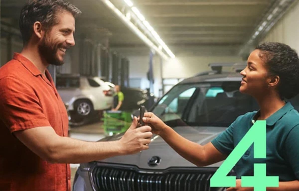 Ein Mann erhält in einer Werkstatt von einer Mitarbeiterin den Autoschlüssel überreicht, während beide sich anlächeln und ein Fahrzeug im Hintergrund steht.