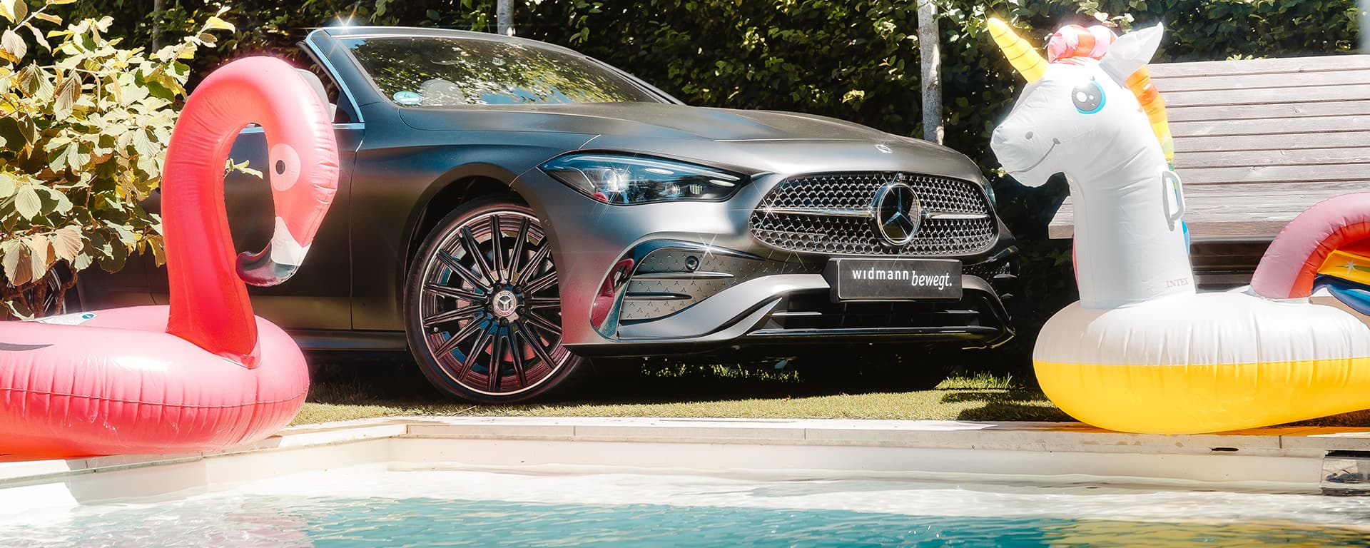 Ein aufblasbarer pinker Flamingo-Schwimmreif und ein aufblasbarer weißer Einhorn-Schwimmreif in einem Swimming Pool im Vordergrund. Im Hintergrund ein mattgraues Mercedes-Benz Cabrio.