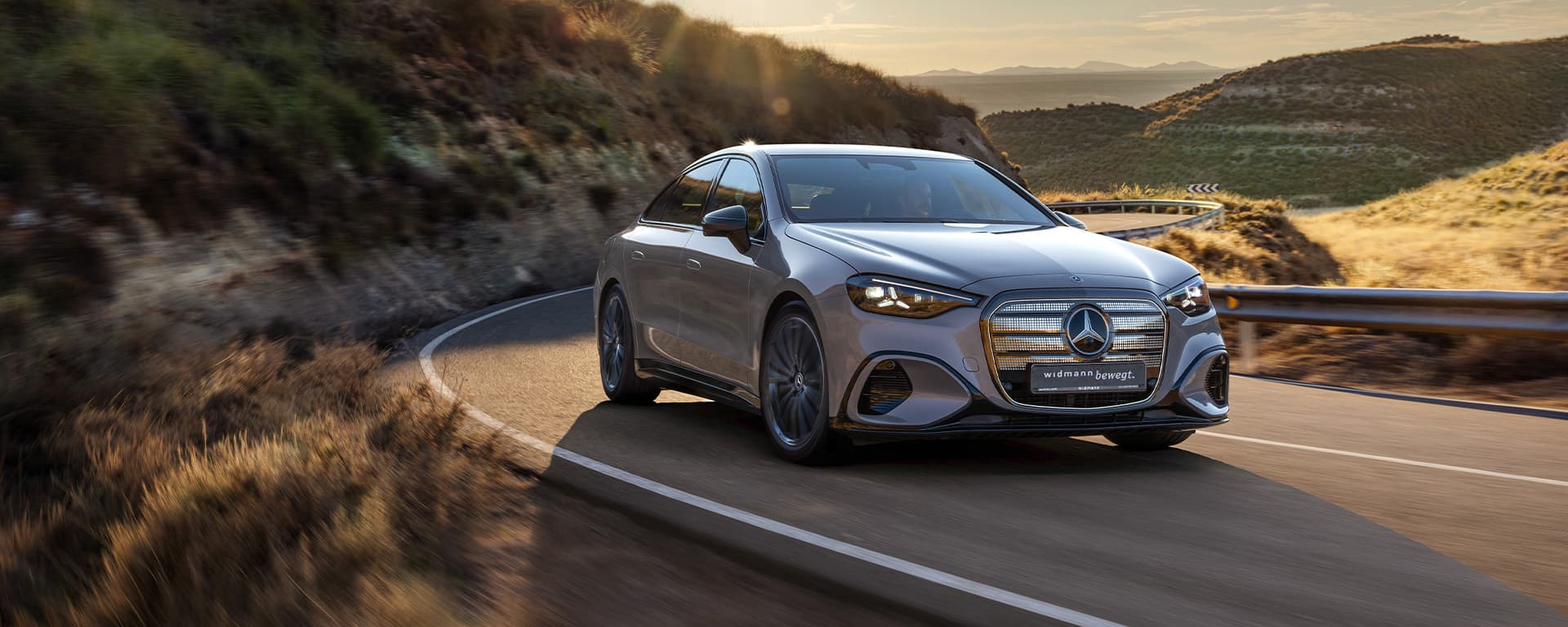Ein silbernes Auto fährt auf einer kurvigen Straße durch eine hügelige Landschaft bei tief stehender Sonne.