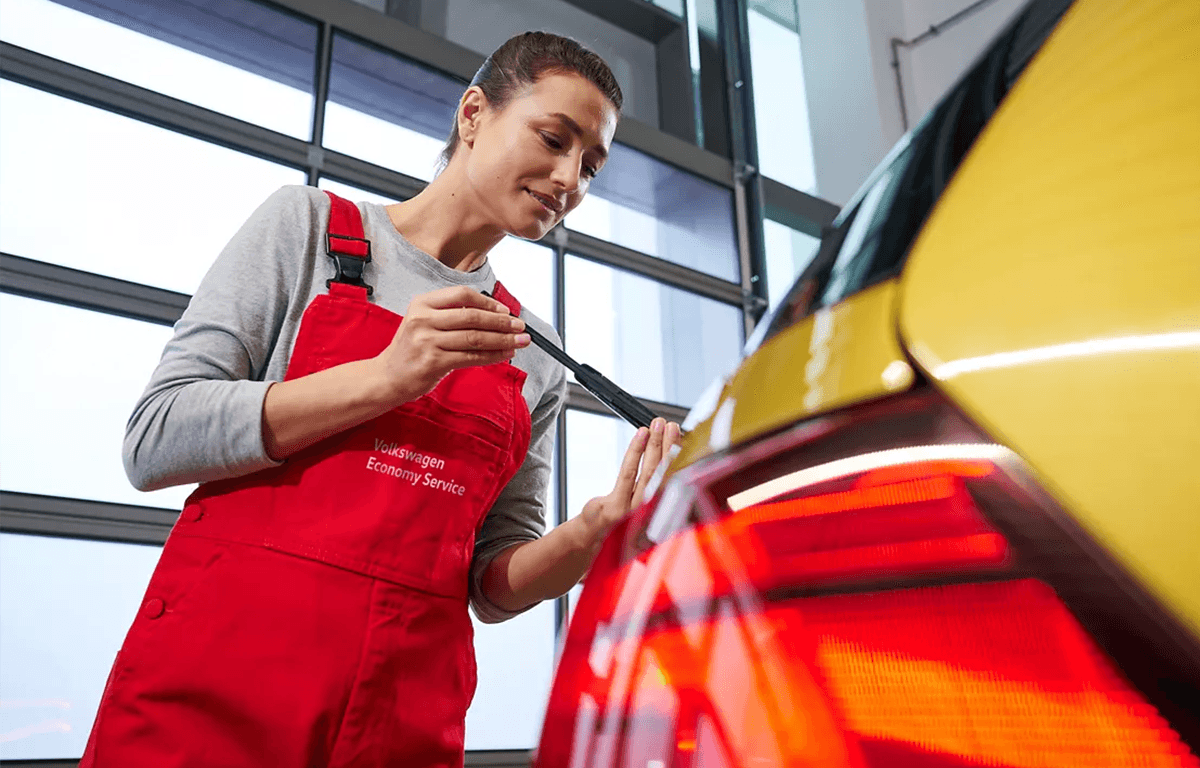 Person in roter Arbeitskleidung prüft die Heckleuchte eines gelben Autos.