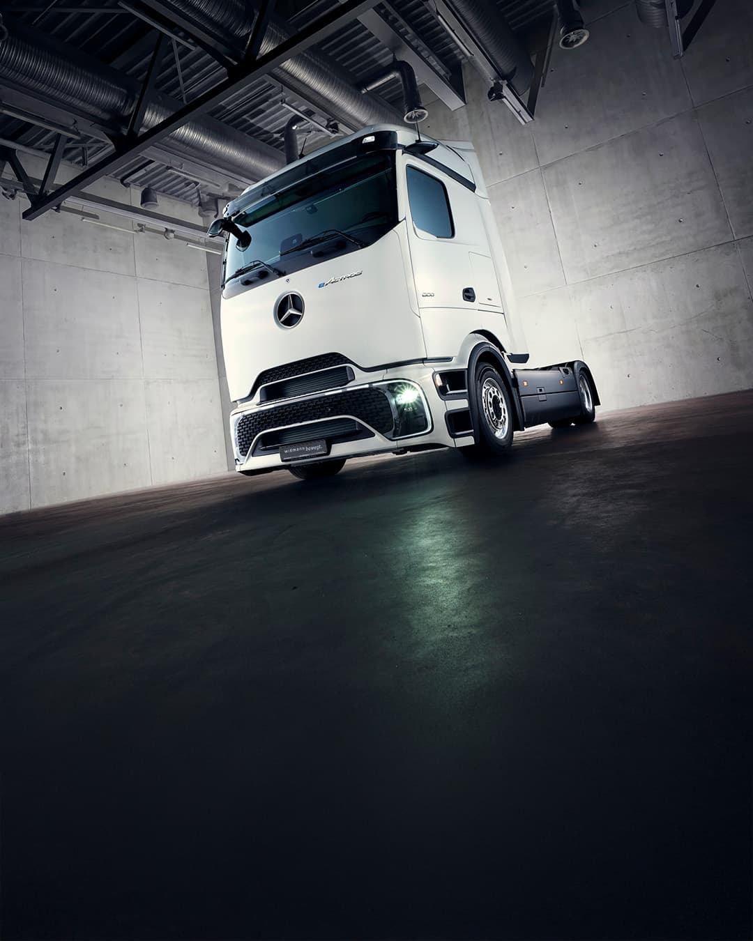 Ein weißer Mercedes-Benz Lkw steht in einer modernen, industriellen Halle mit Betonwänden und dunklem Boden, eindrucksvoll beleuchtet durch einen hellen Lichtkegel.