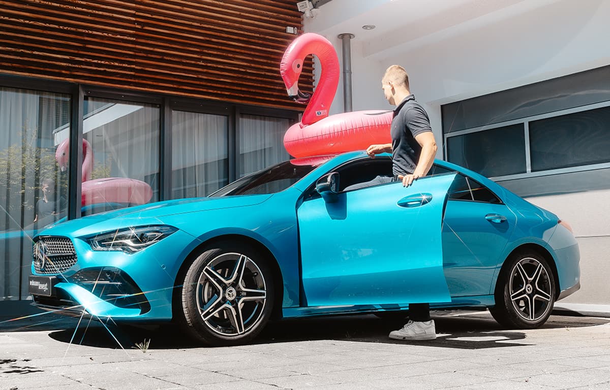 Ein Mann steigt in einen blauen Mercedes-Benz CLA ein, auf dessen Dach ein pinker, aufblasbarer Flamingo-Schwimmreif liegt.