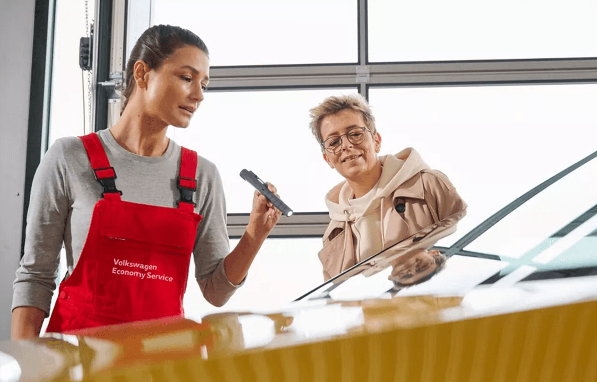 Zwei Personen in einer Werkstatt begutachten gemeinsam die Frontscheibe eines Autos, eine trägt Arbeitskleidung mit „Volkswagen Economy Service“.