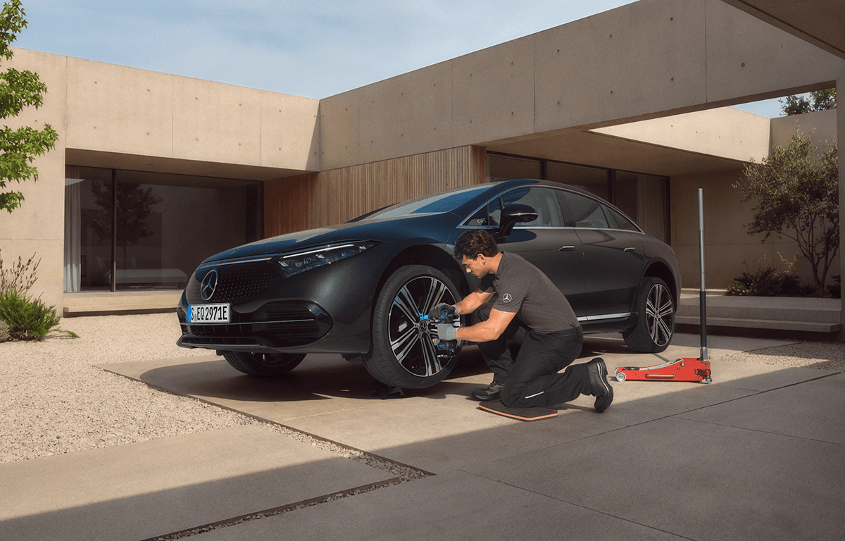 Ein Techniker kniet vor einem schwarzen Auto und führt vor einem modernen Gebäude eine Wartung am Fahrzeug durch.