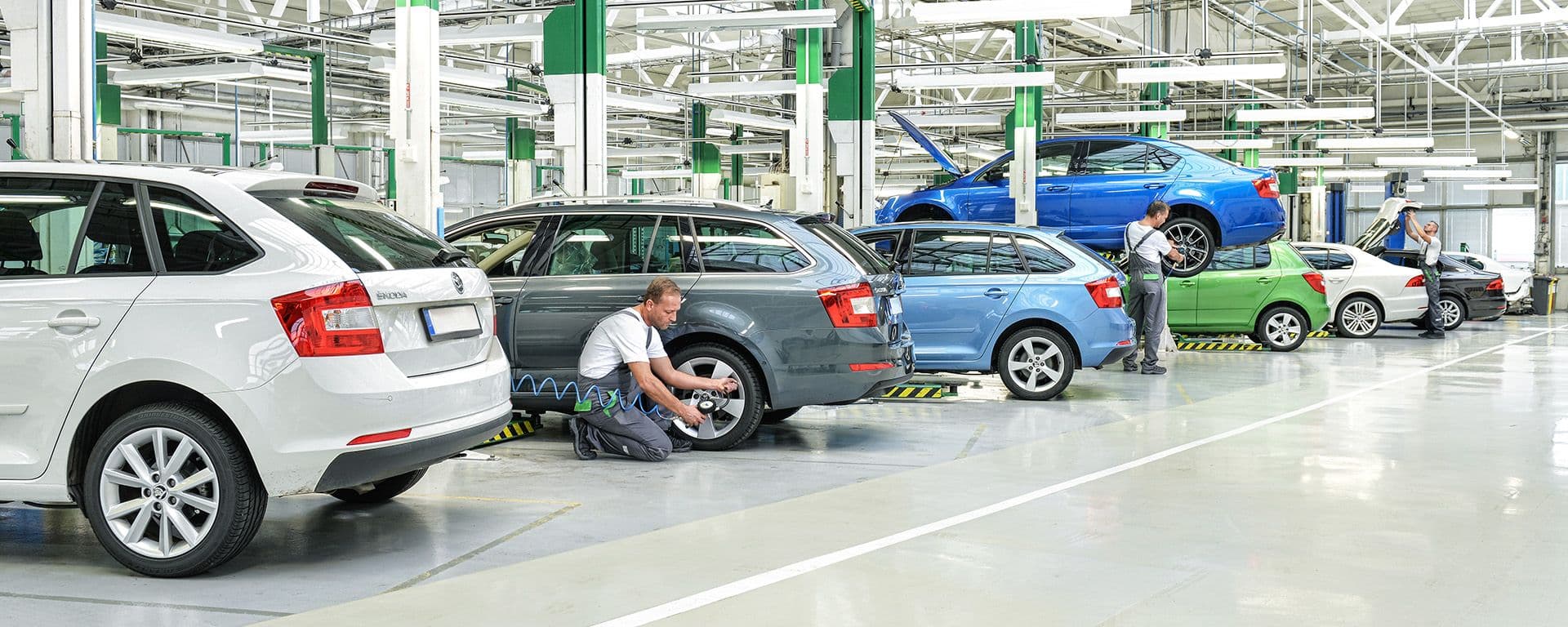Mehrere Fahrzeuge stehen in einer großen Werkstatt, während ein Mechaniker an einem Auto arbeitet und andere Fahrzeuge auf Hebebühnen gewartet werden