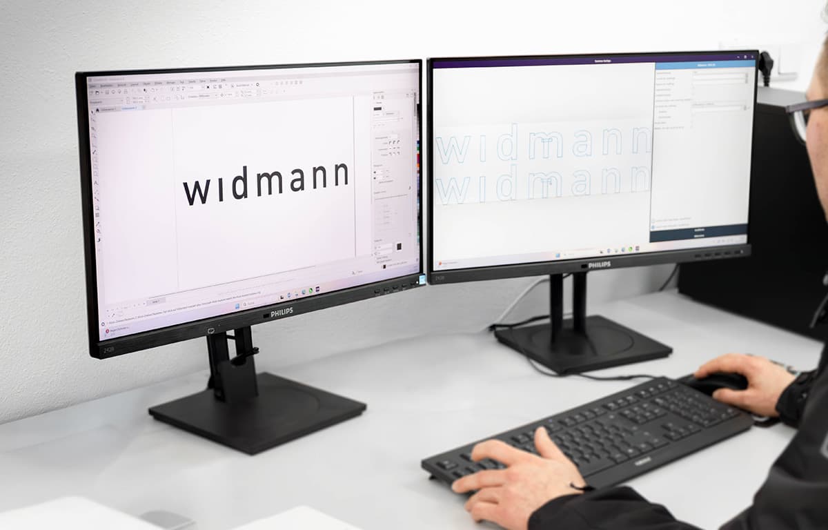 Eine Person arbeitet an einem Schreibtisch mit zwei Monitoren, auf denen das Wort „widmann“ in einem Grafikprogramm gestaltet wird.