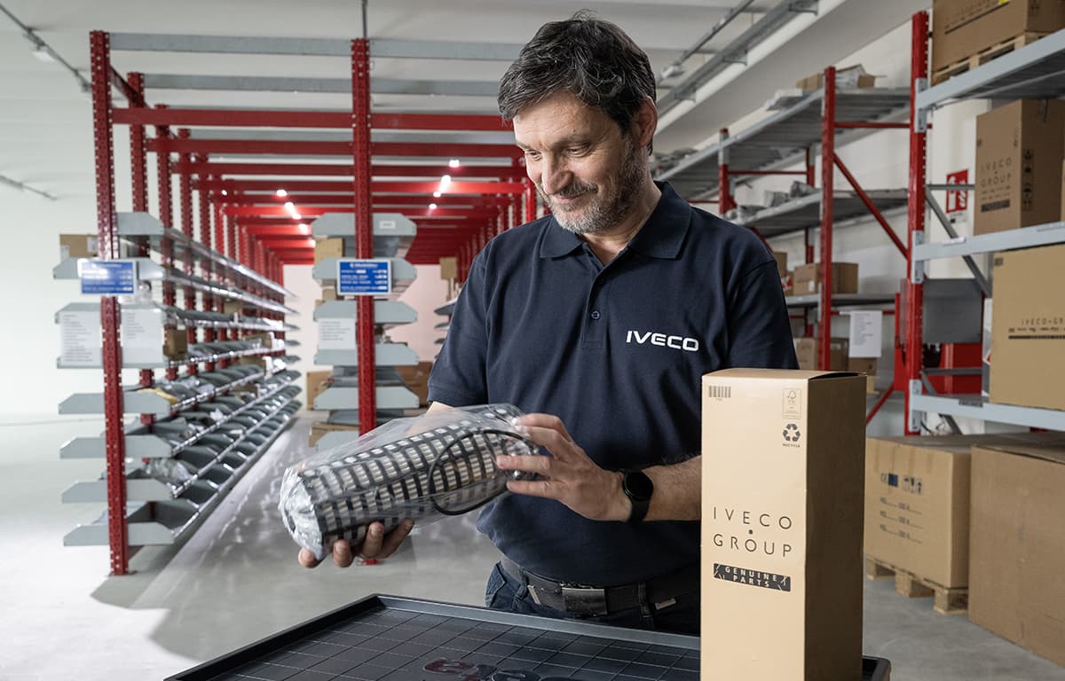 Ein Mitarbeiter steht in einem Lager und hält ein ausgepacktes Ersatzteil in der Hand.