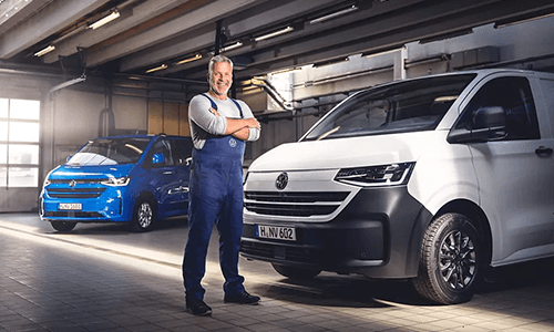 Ein Mitarbeiter in Arbeitskleidung steht in einer Werkstatt neben einem weißen Volkswagen Transporter und einem blauen Fahrzeug.