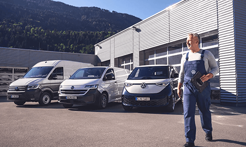 Ein Mitarbeiter in Arbeitskleidung steht vor einem Werkstattgebäude neben mehreren geparkten Volkswagen Nutzfahrzeugen.