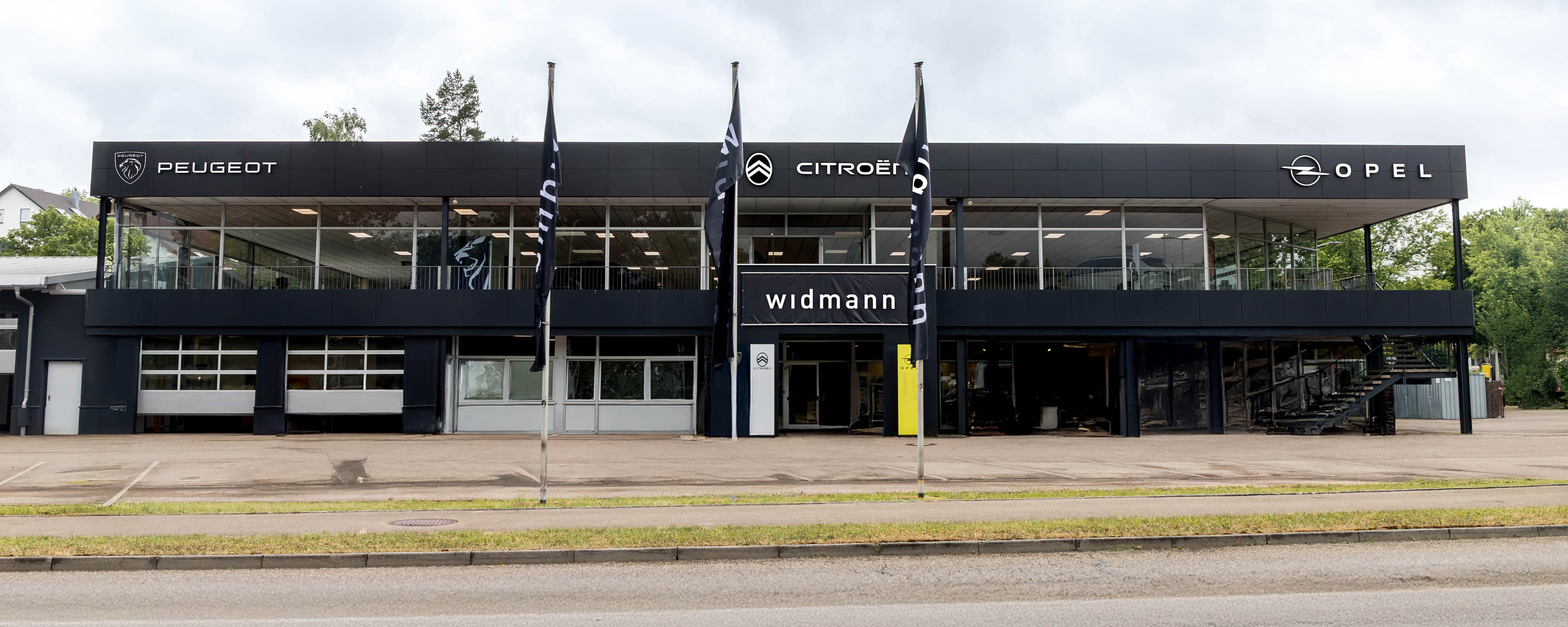 Außenansicht des Autohauses Widmann mit den Marken Peugeot, Citroën und Opel sowie modernem Showroom an einer Straße.