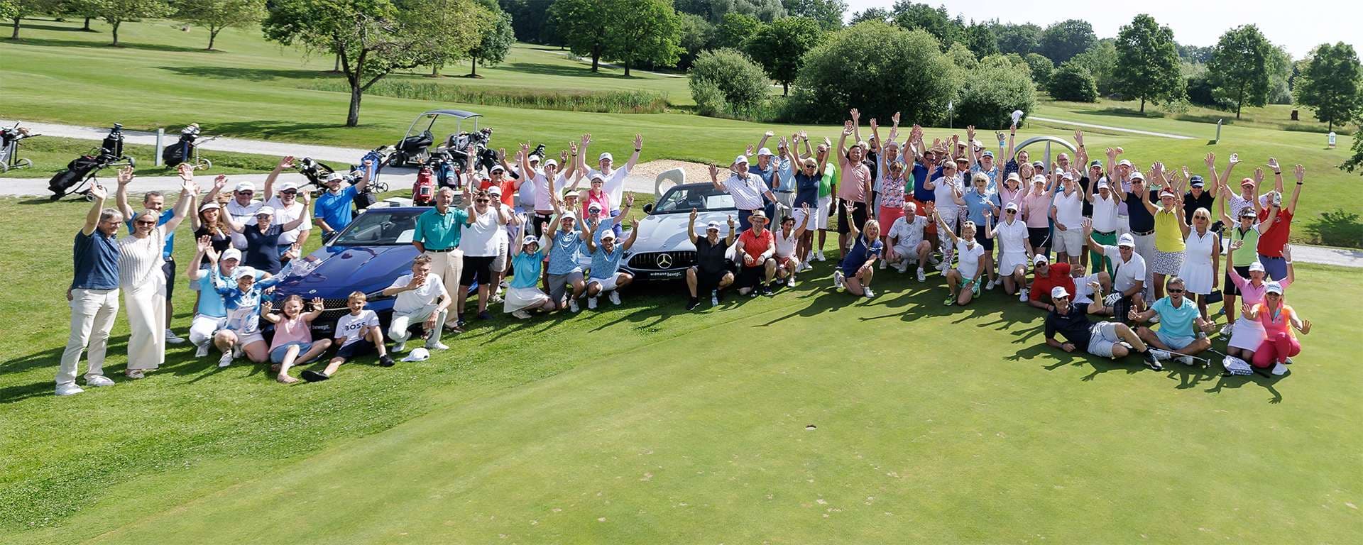 Die Teilnehmer eines Golfturniers in einem Halbkreis auf einem Golfplatz. Viele strecken die Arme in die Luft.