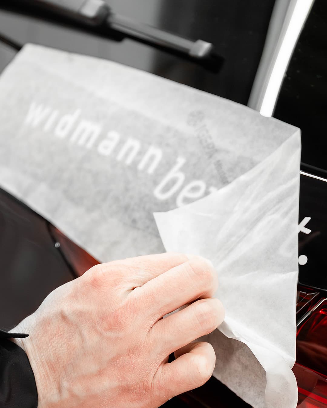Eine Person zieht eine transparente Schutzfolie mit dem Schriftzug „Widmann bewegt“ von der Heckscheibe eines schwarzen Autos ab.