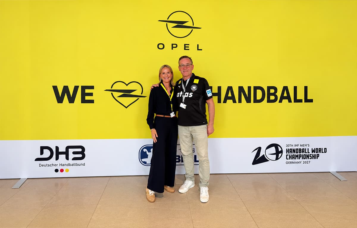 Zwei Personen stehen gemeinsam vor einer gelben Opel-Fotowand mit der Aufschrift „WE LOVE HANDBALL“ und dem Logo der Handball-Weltmeisterschaft 2027.