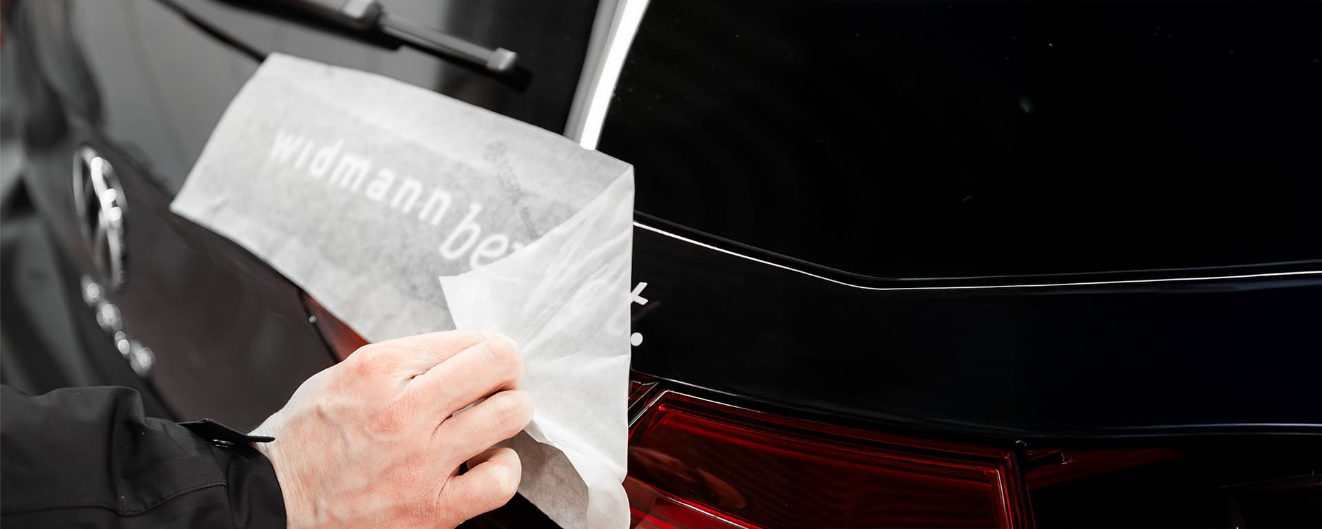 Eine Person zieht eine transparente Schutzfolie mit dem Schriftzug „Widmann bewegt“ von der Heckscheibe eines schwarzen Autos ab.